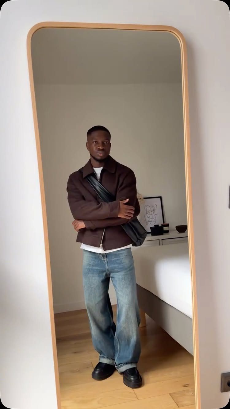 Man wearing a brown jacket, wide-leg blue jeans, and black leather derby shoes.