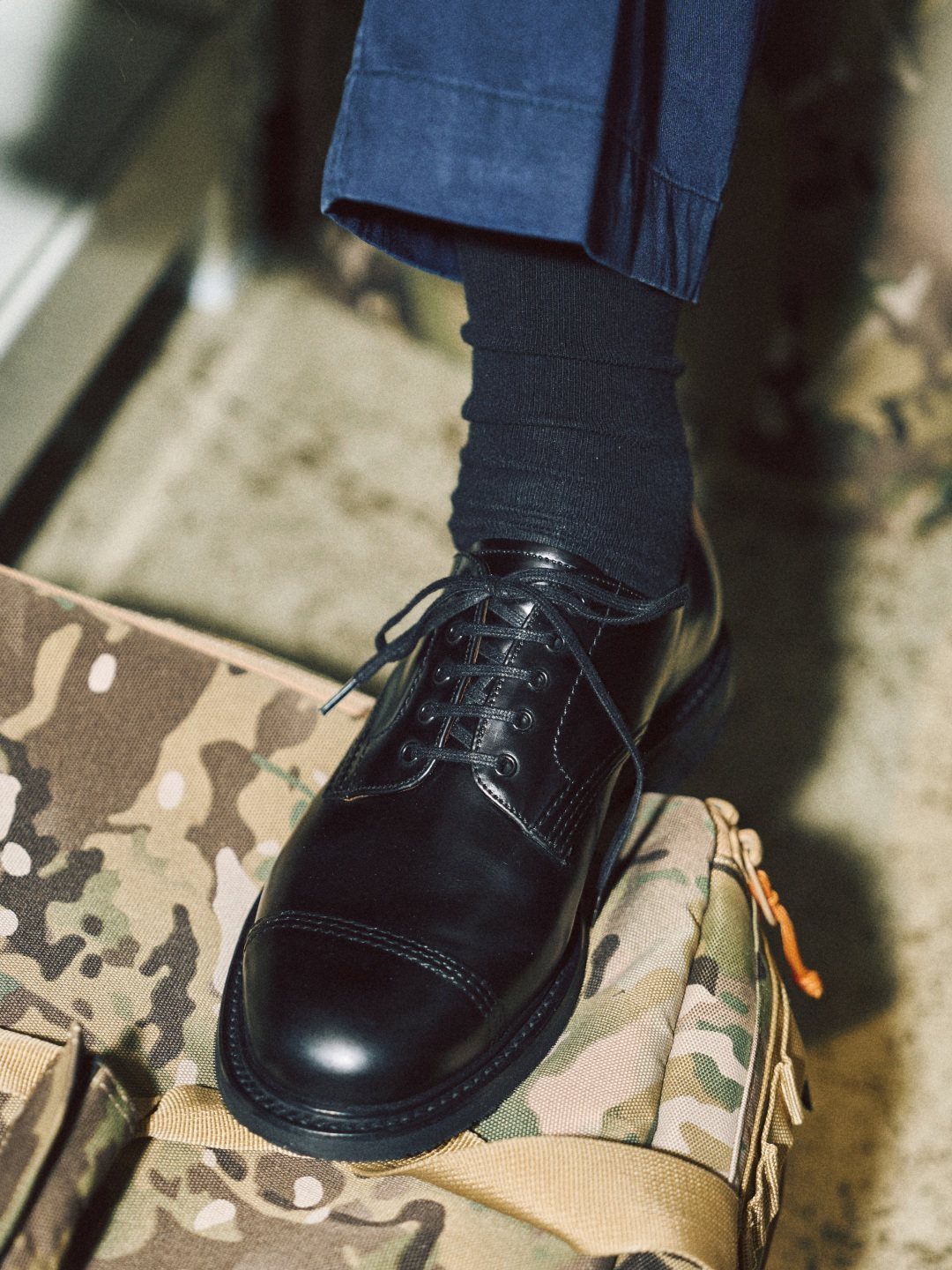 Black leather cap-toe derby shoe worn with blue trousers and black socks.