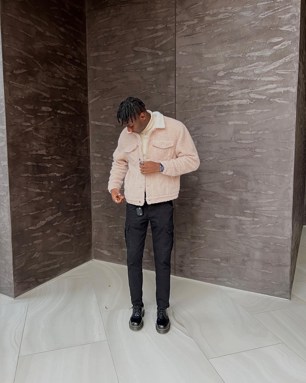 Person in a beige fleece jacket and black trousers standing by a marbled wall.