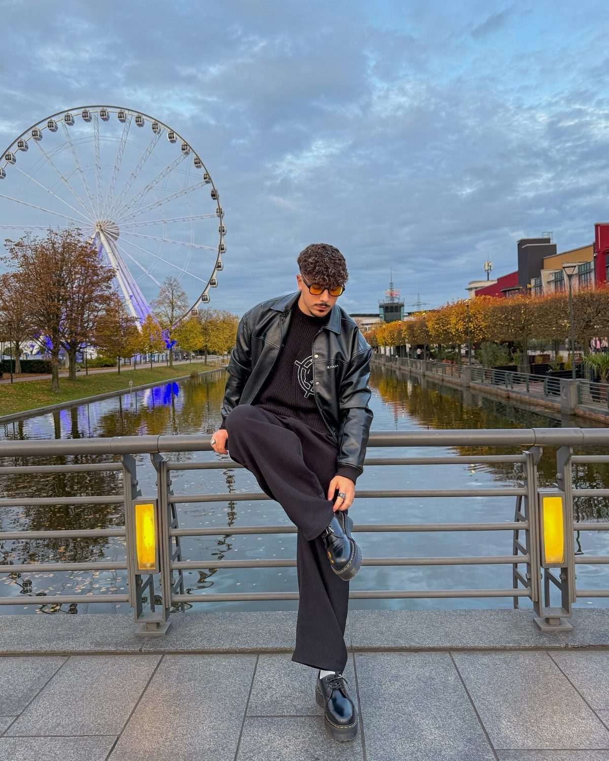 Man in black leather jacket and wide-leg trousers posing by a riverfront Ferris wheel.