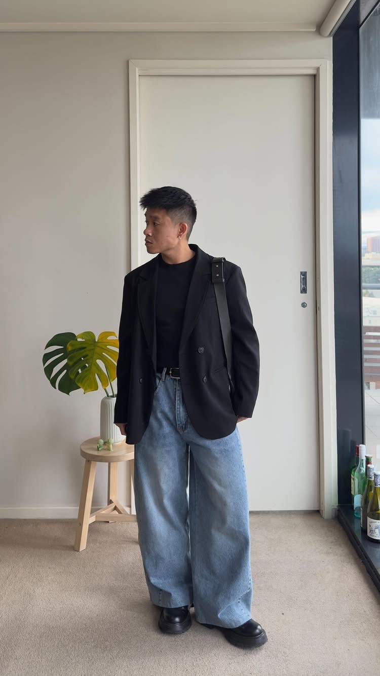 Man wearing a black oversized blazer and wide-leg light blue jeans indoors.