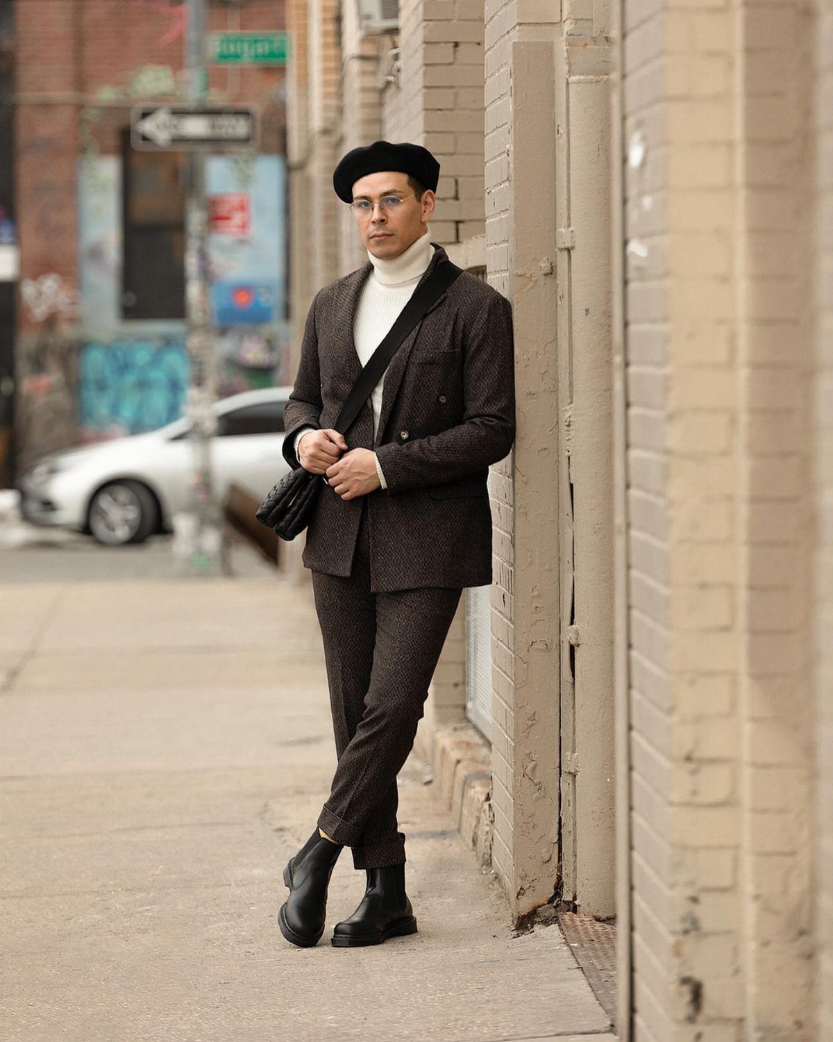 Man in a brown suit and white turtleneck leaning against a brick wall.