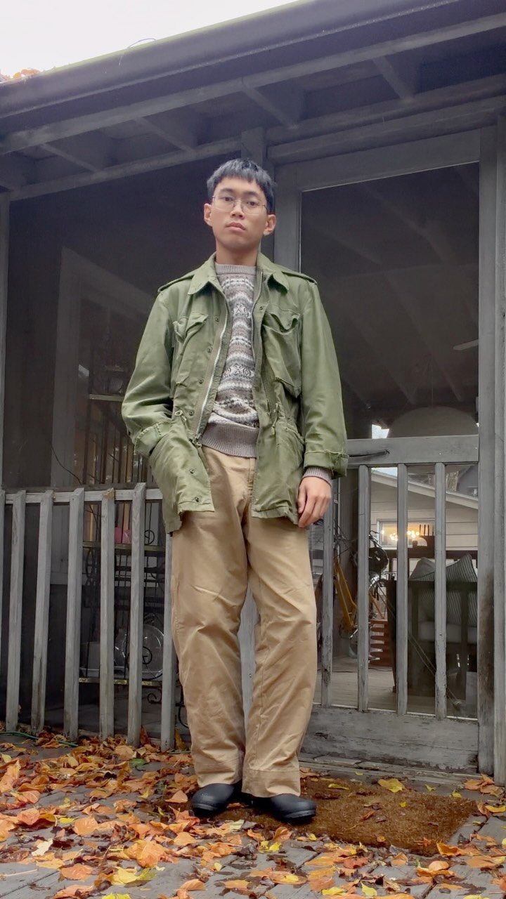 Person in olive green utility jacket and tan trousers standing on leaf-covered porch.