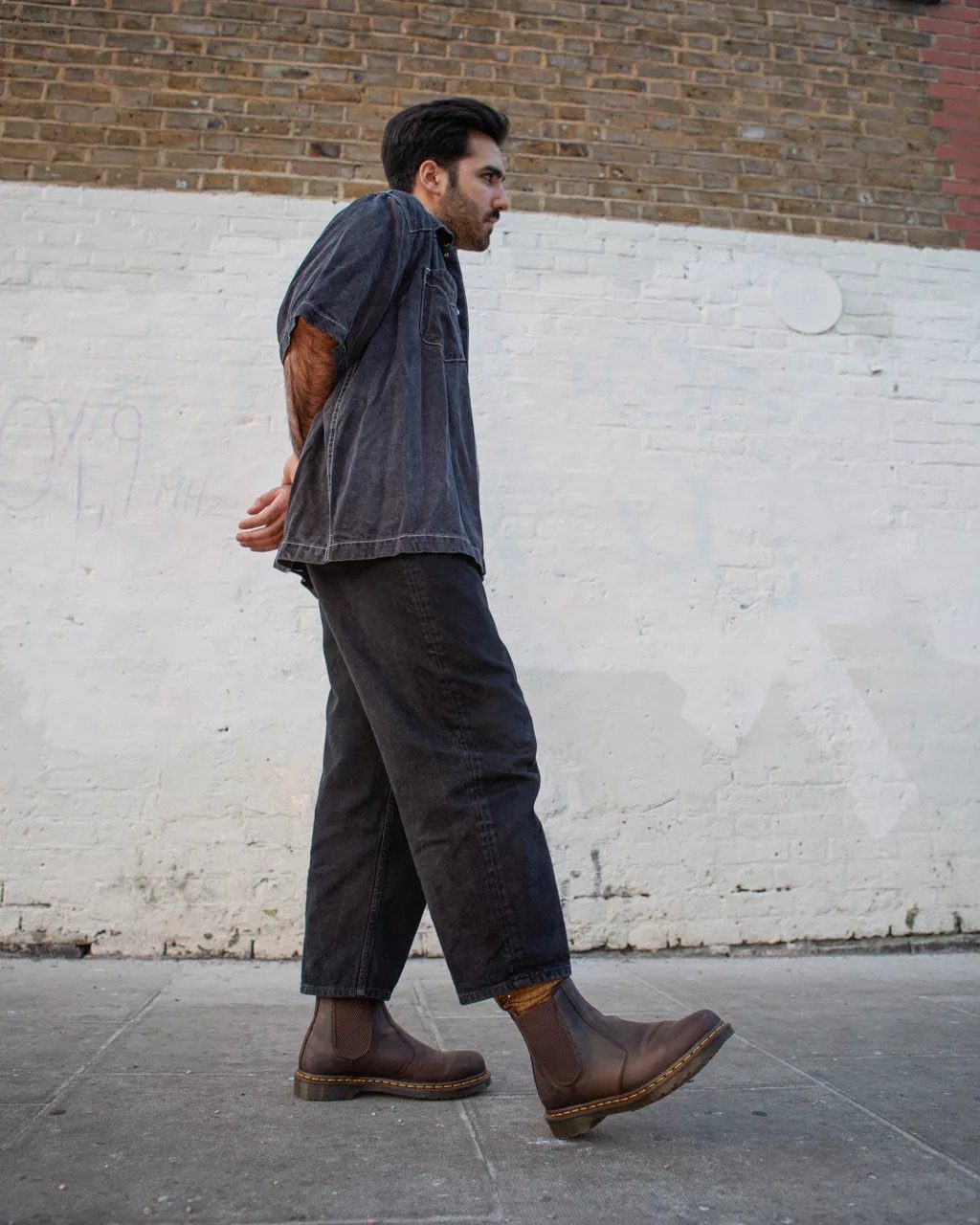Man wearing charcoal trousers and brown leather Chelsea boots standing by a white wall.