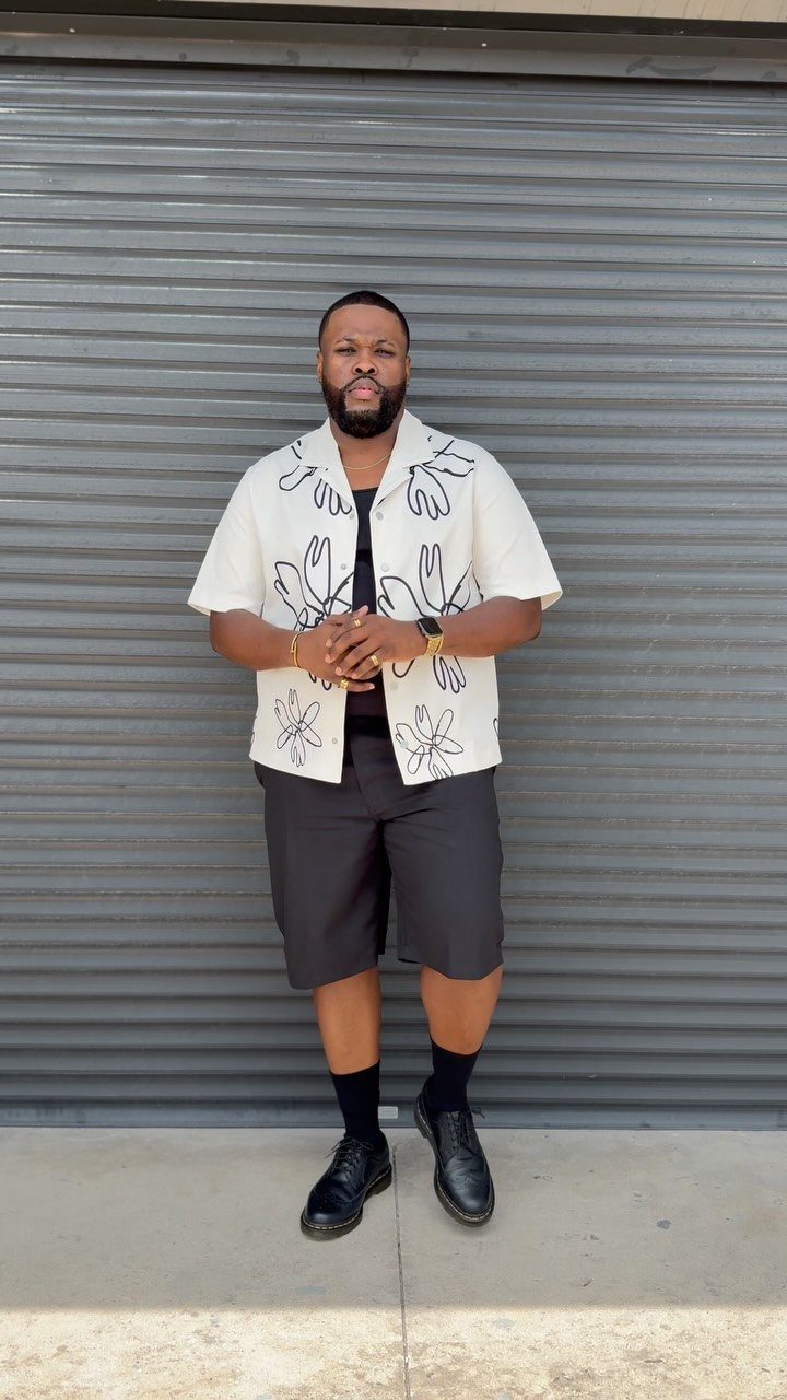 Man wearing a floral white shirt, grey shorts, and black Dr Martens shoes.