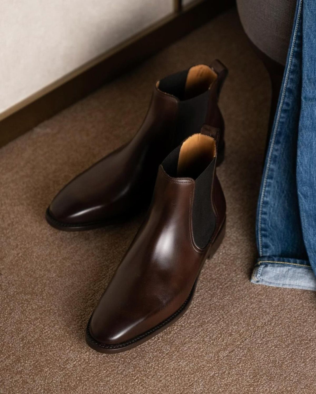 Dark brown leather Chelsea boots on a brown carpet next to blue jeans.