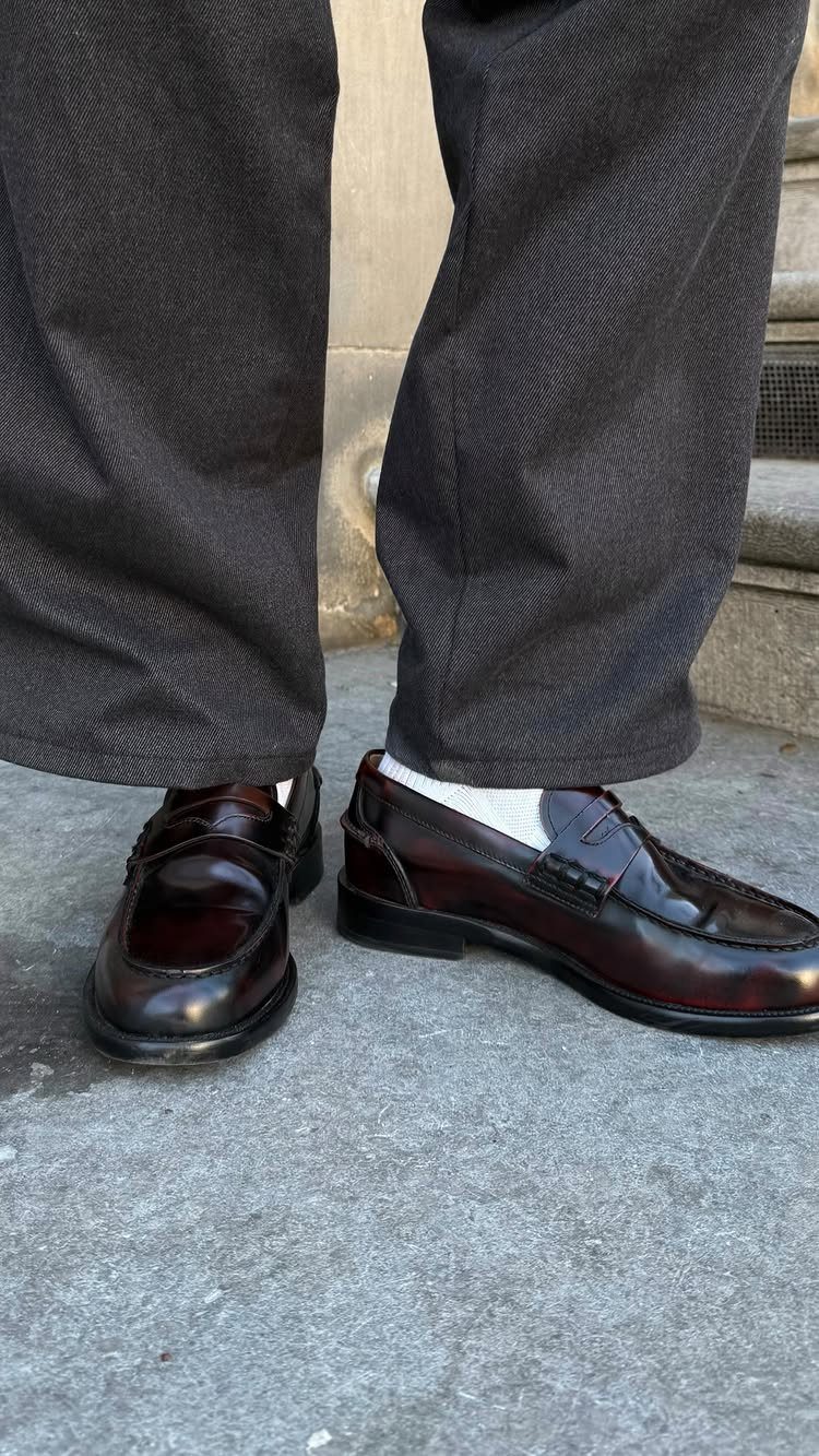Burgundy leather penny loafers with white socks and dark grey trousers on pavement.