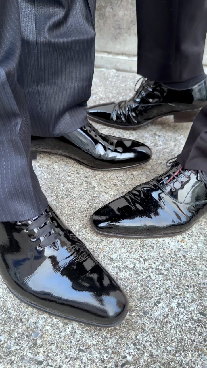 Black patent leather dress shoes worn with dark trousers on a concrete ground.