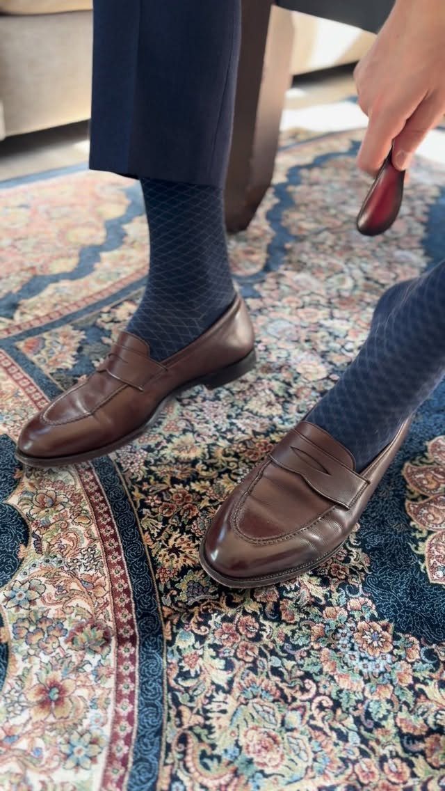 Brown leather penny loafers worn with navy socks and trousers on a patterned rug.