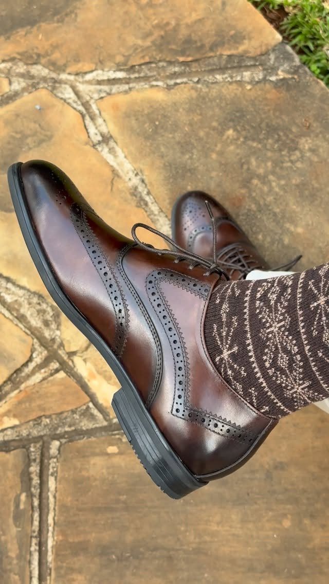 Brown leather wingtip oxford shoes with patterned socks on a stone surface.