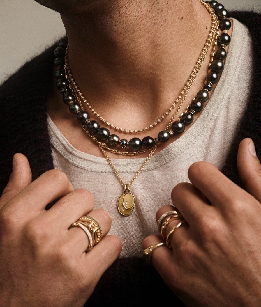 Man wearing layered gold chains and dark pearl necklaces with a coin pendant