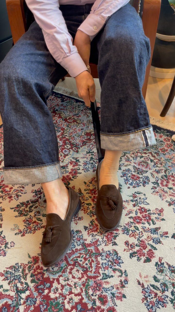 Dark blue cuffed jeans and brown suede tassel loafers on a patterned rug.