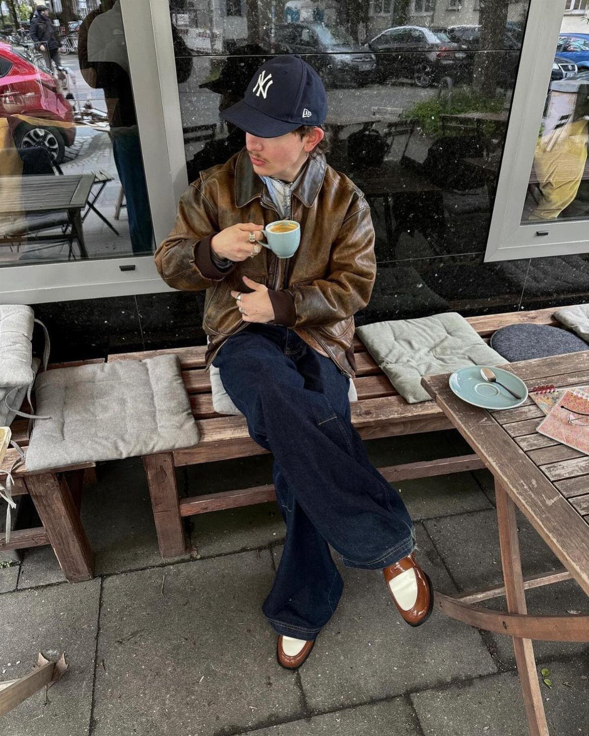 Man wearing brown leather jacket, dark blue jeans, and brown loafers sitting outdoors.