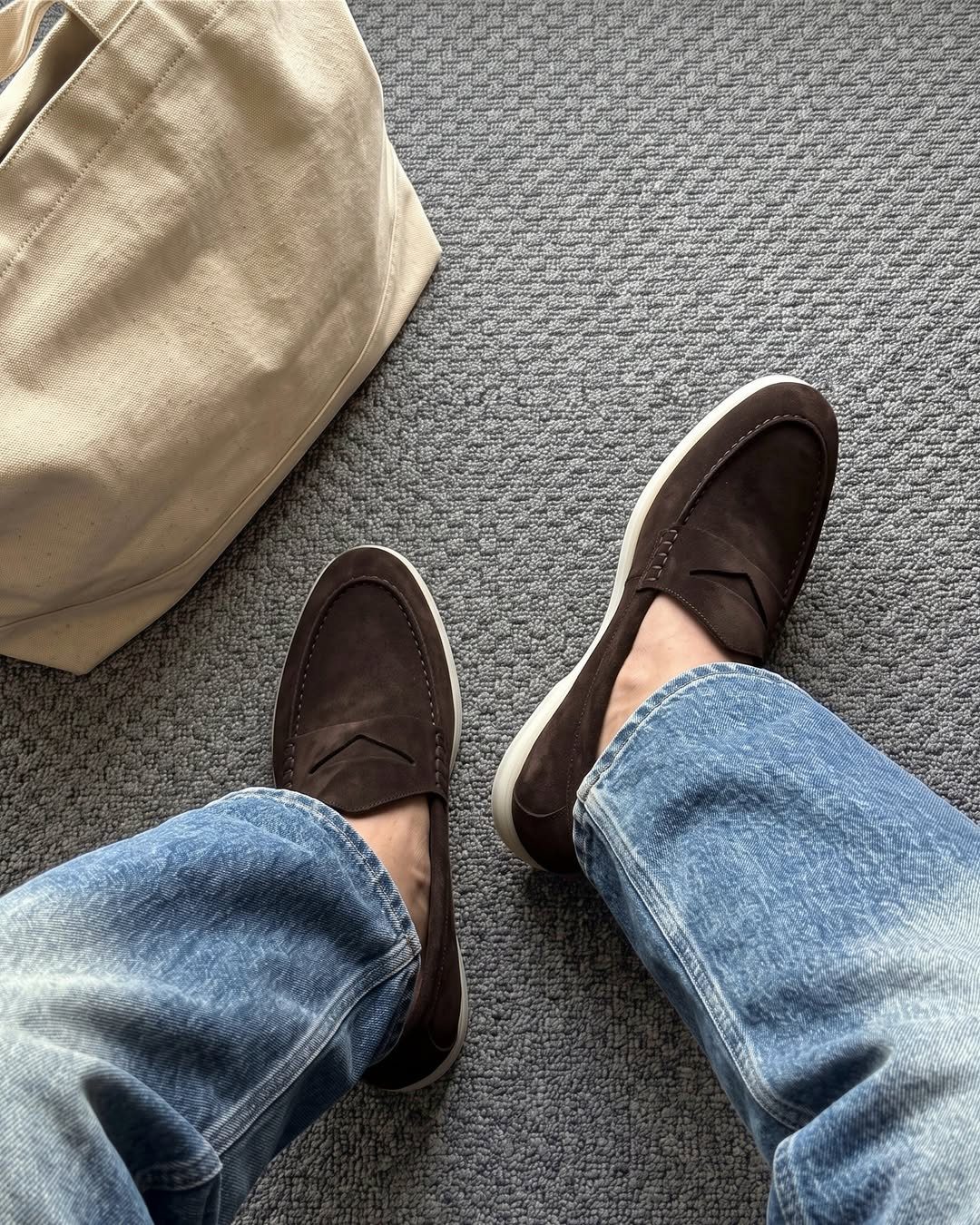 Brown suede loafers and blue jeans on a grey carpet with a beige bag.