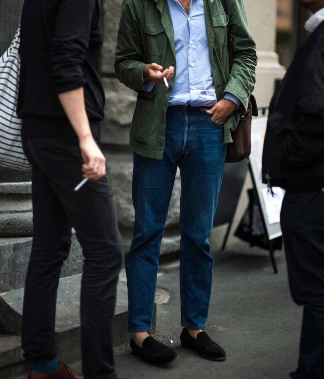 Man wearing a green jacket, blue jeans, and black loafers standing on a street
