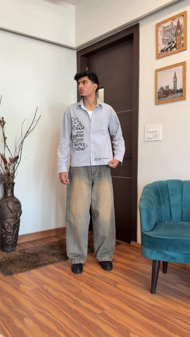 Man wearing a grey shirt, wide-leg faded jeans, and black loafers indoors.