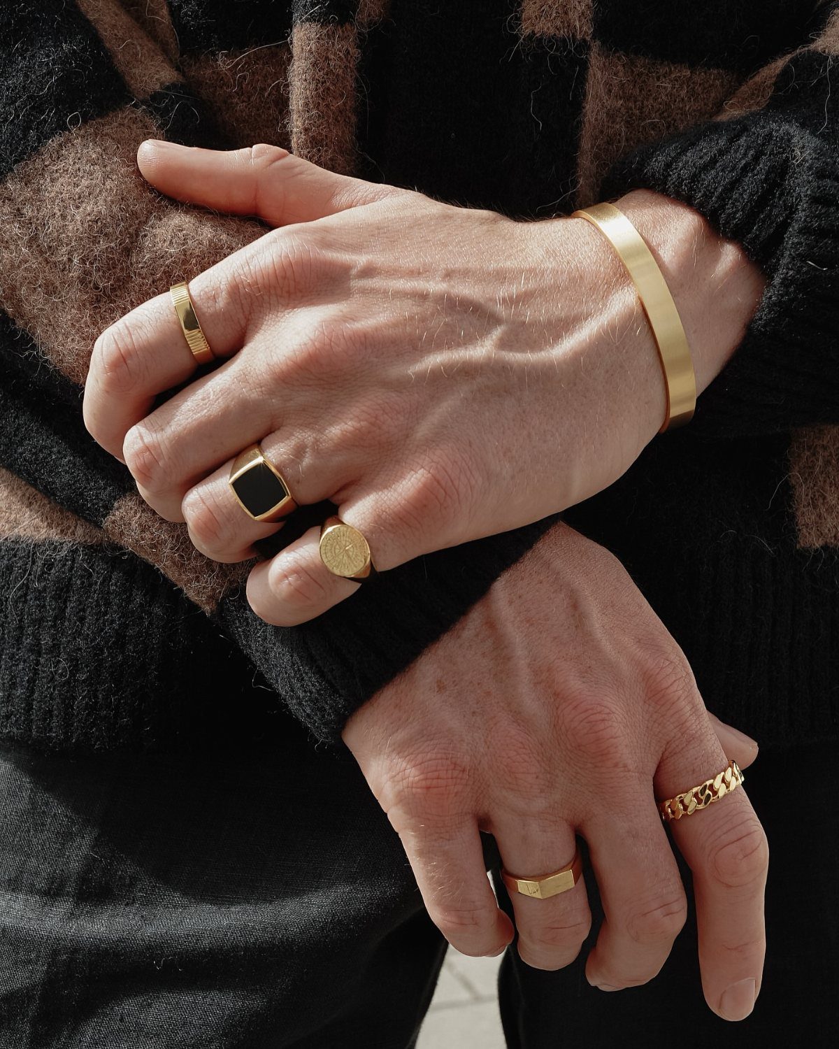 Hands wearing gold signet rings and a gold cuff bracelet with checkered sweater