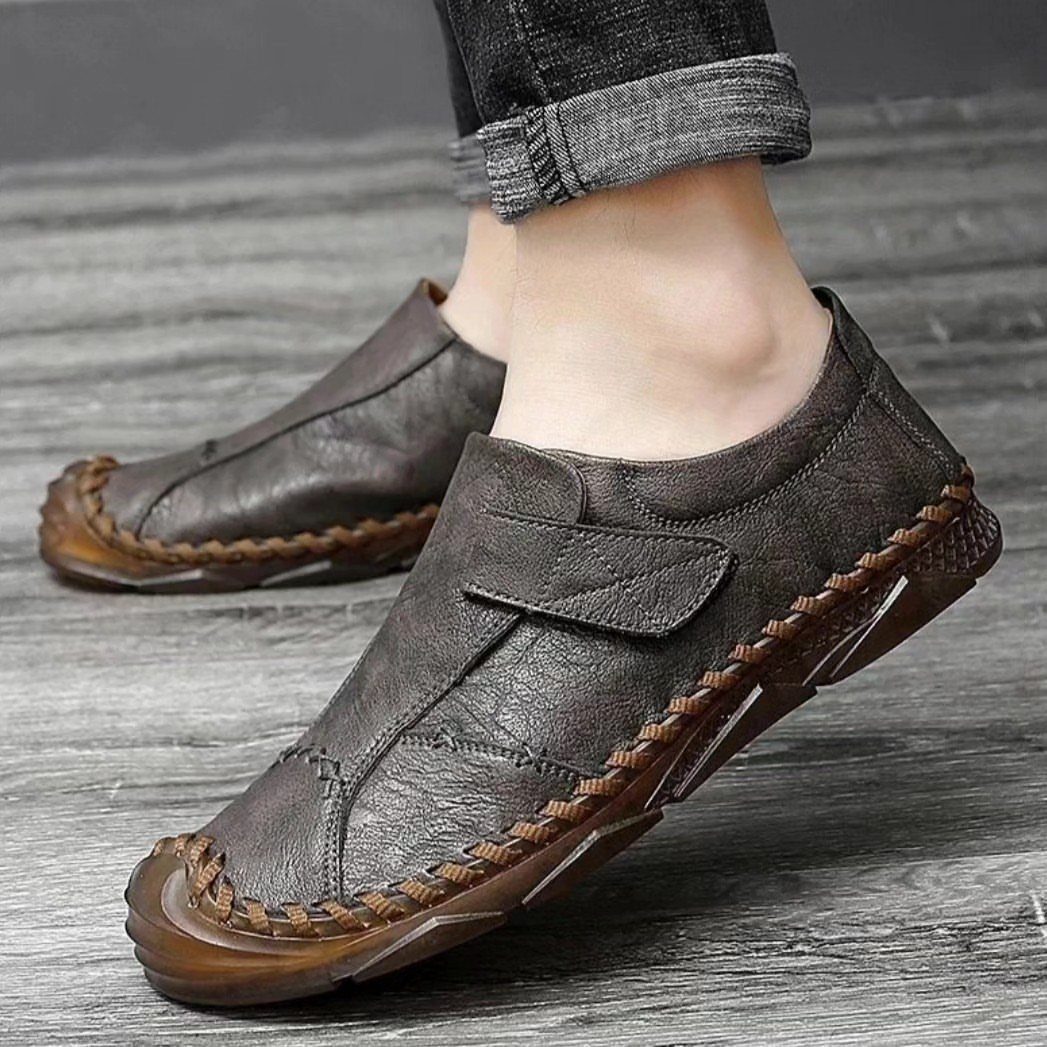 Dark brown leather slip-on shoes with decorative stitching and a strap on feet.