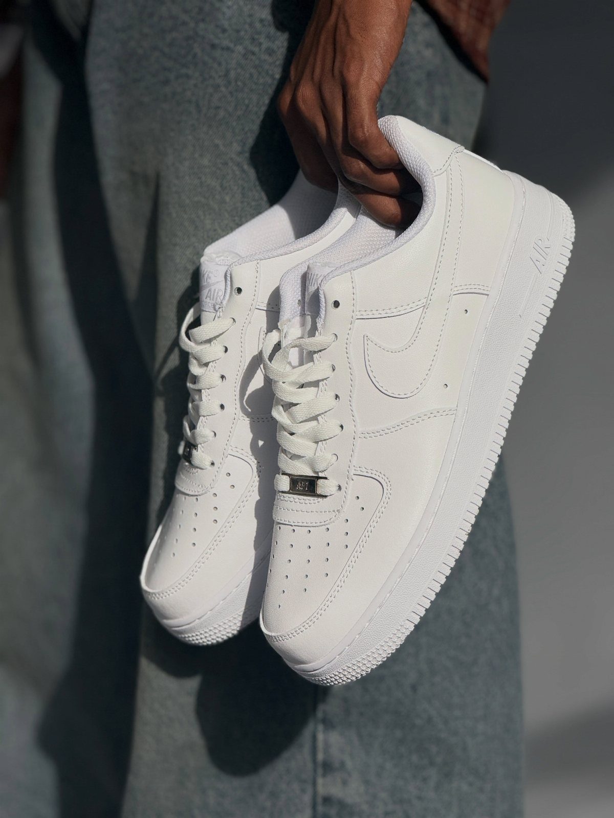 White leather sneakers held by a person wearing greyish-blue trousers against a plain wall.