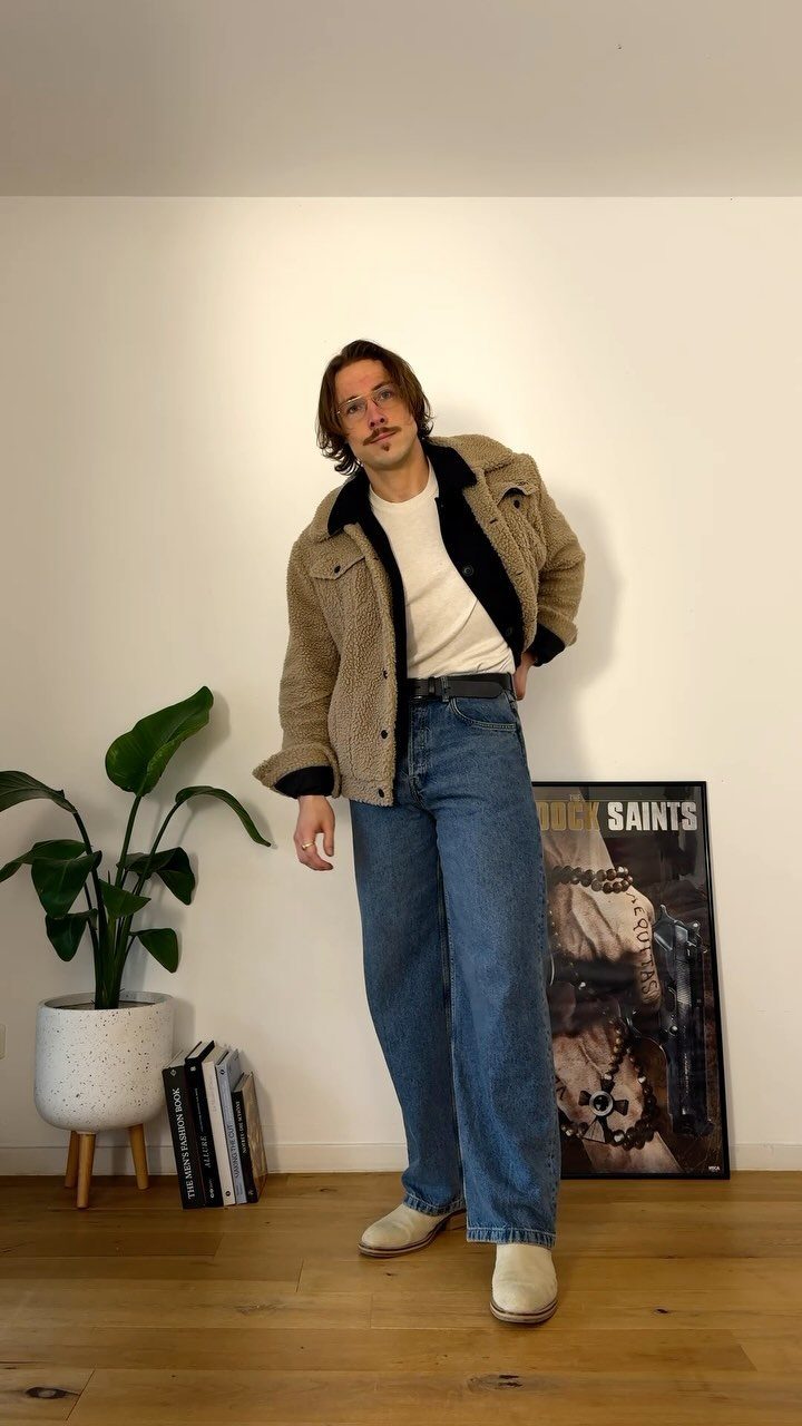 Man wearing a tan sherpa jacket, wide blue jeans, and beige suede boots.