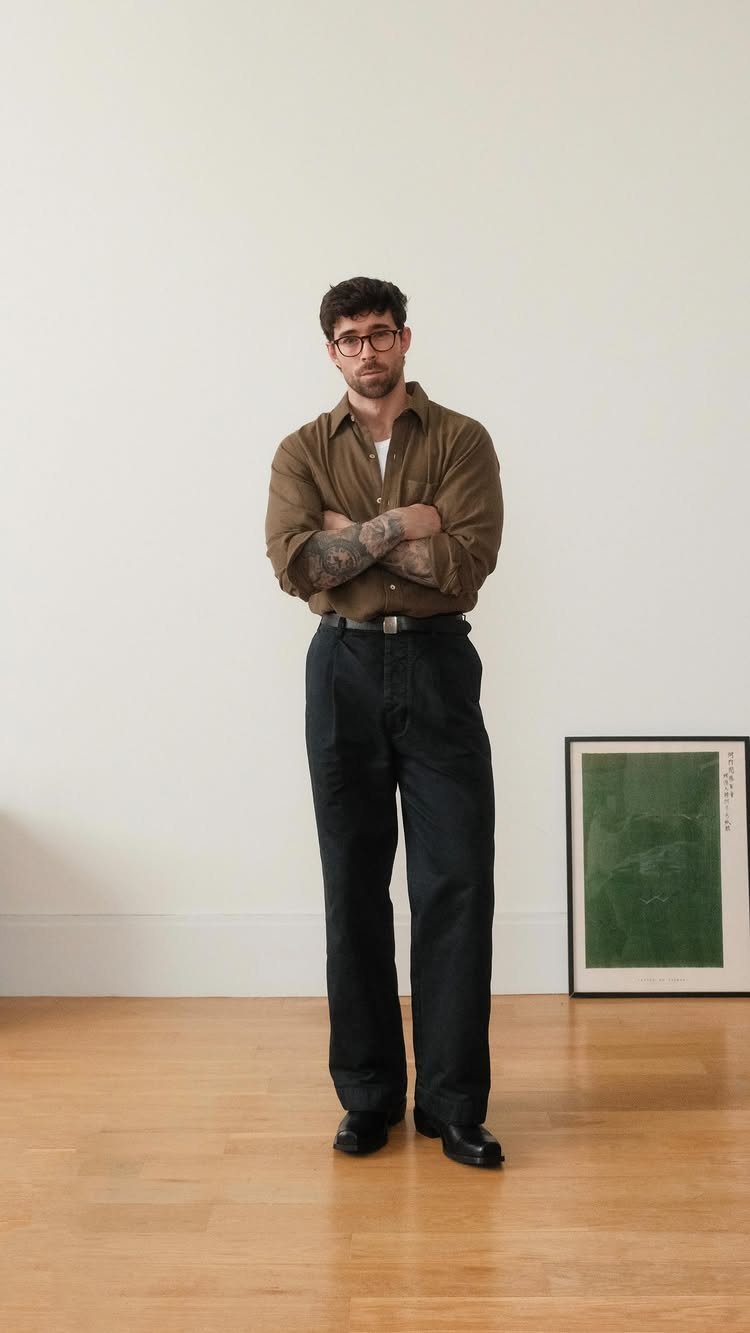 Man wearing a brown shirt, black trousers, and black leather boots standing indoors.
