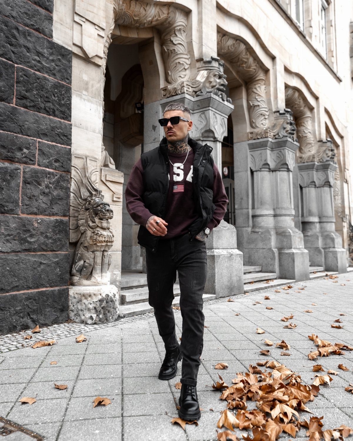 Man in black leather boots, black pants, and maroon sweatshirt standing on city sidewalk