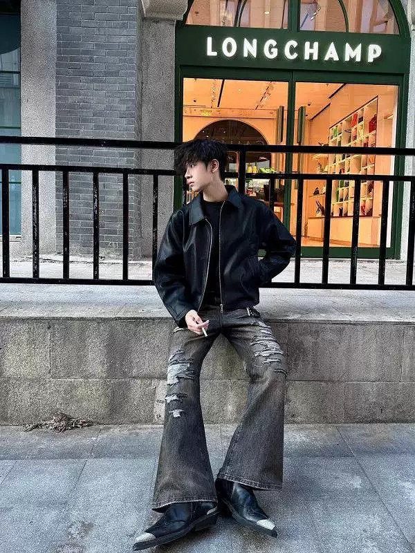 Person in black leather jacket and pointed boots sitting outside a store.