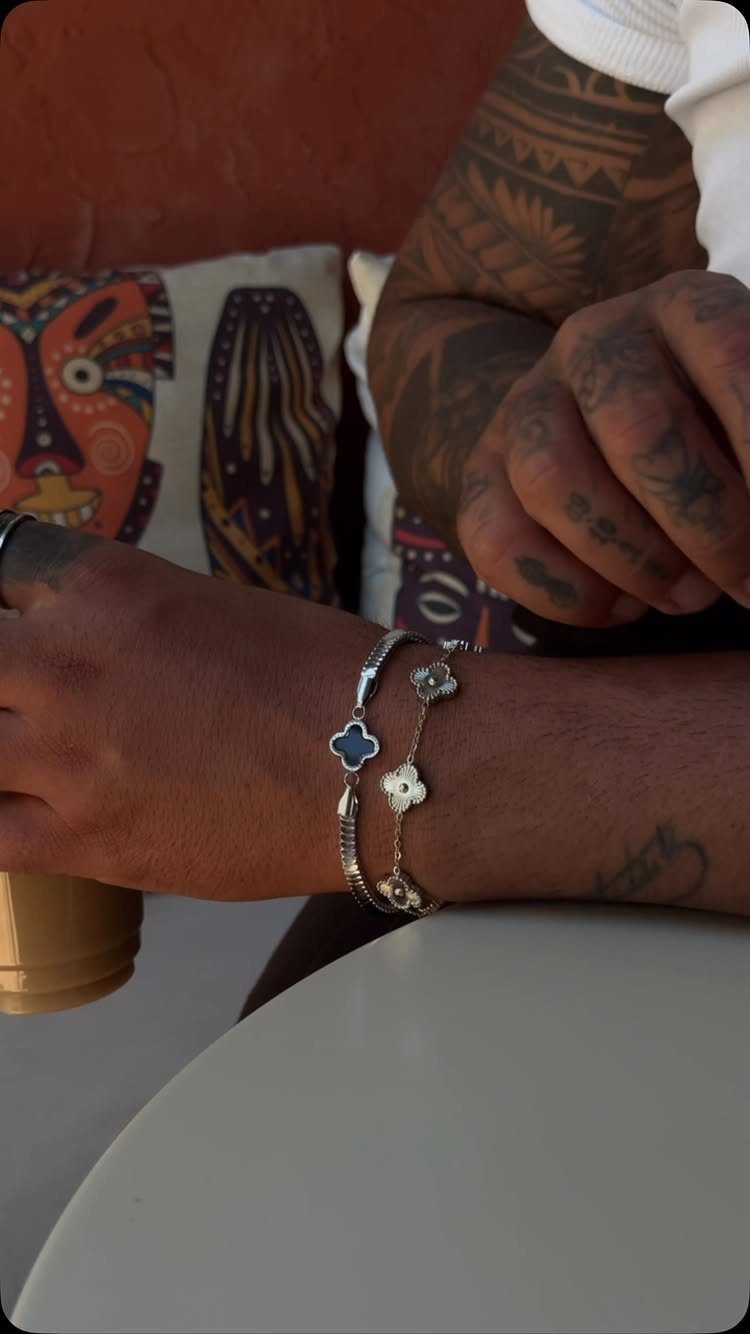 Tattooed wrist wearing a stack of two silver clover bracelets at a table