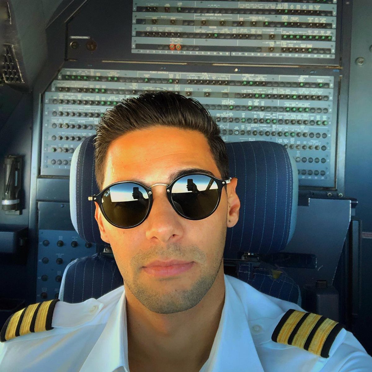 Pilot in white uniform wearing dark aviator sunglasses inside an airplane cockpit.