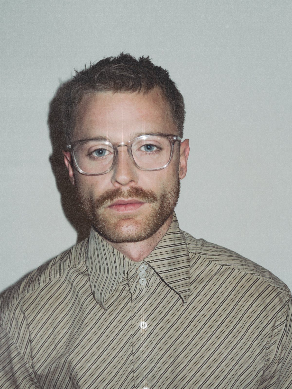 Man with beard wearing clear-framed glasses and a brown striped shirt.
