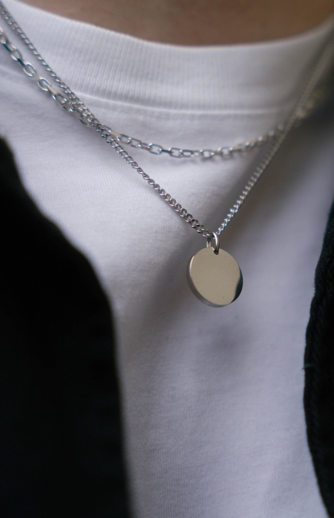 Two silver chain necklaces with a circular pendant worn over a white shirt.