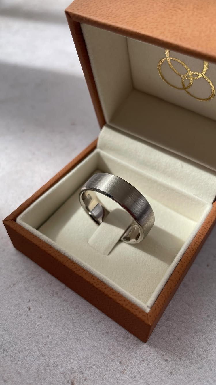 Silver-colored brushed metal ring inside a brown wooden jewelry box with white padding.
