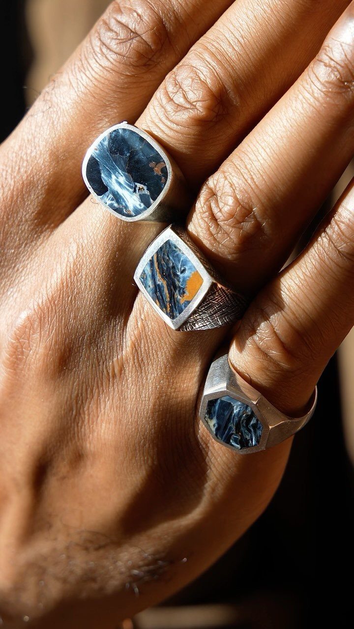 Man's hand wearing three silver rings with blue and orange stone inlays.