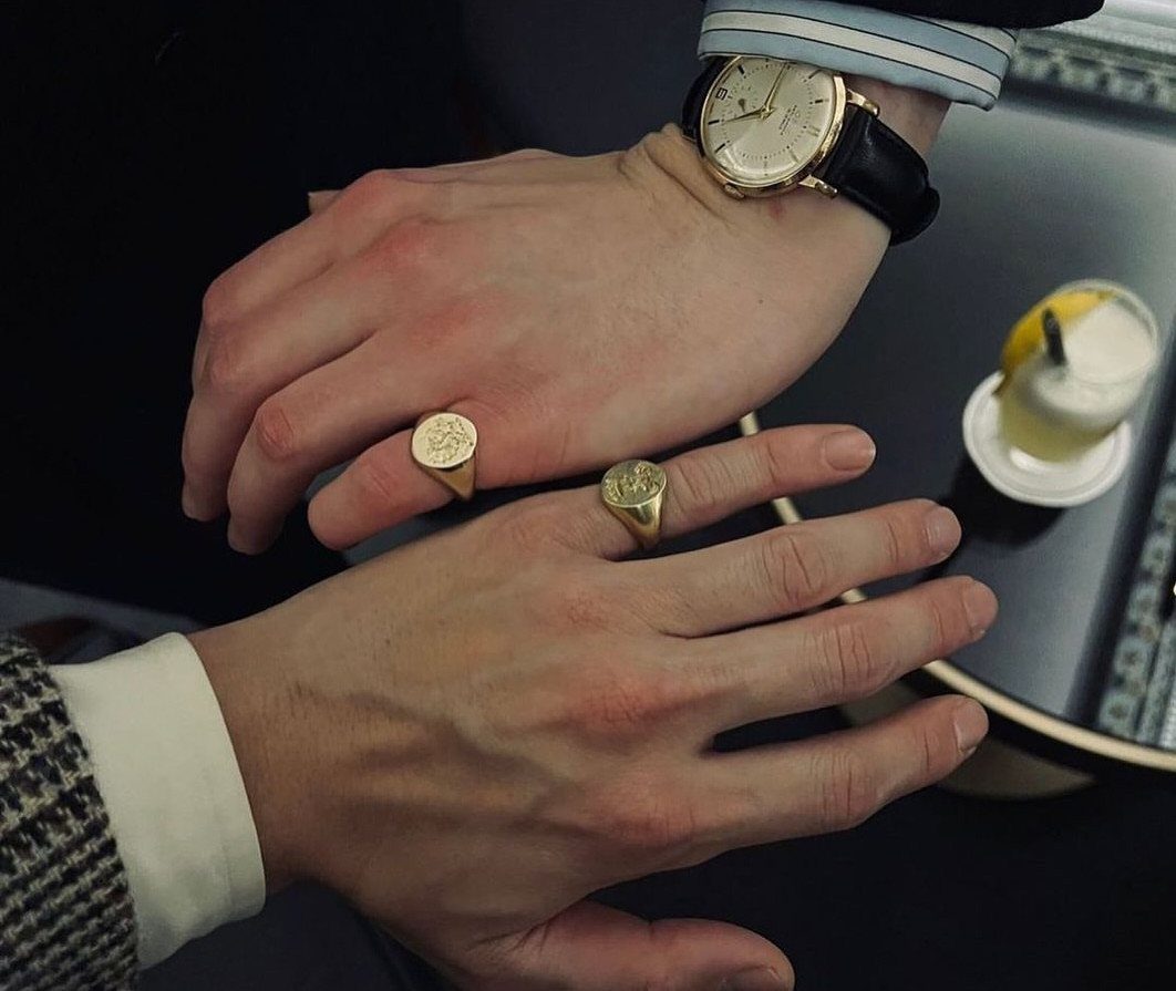 Two men wearing gold signet rings on their fingers next to a watch.