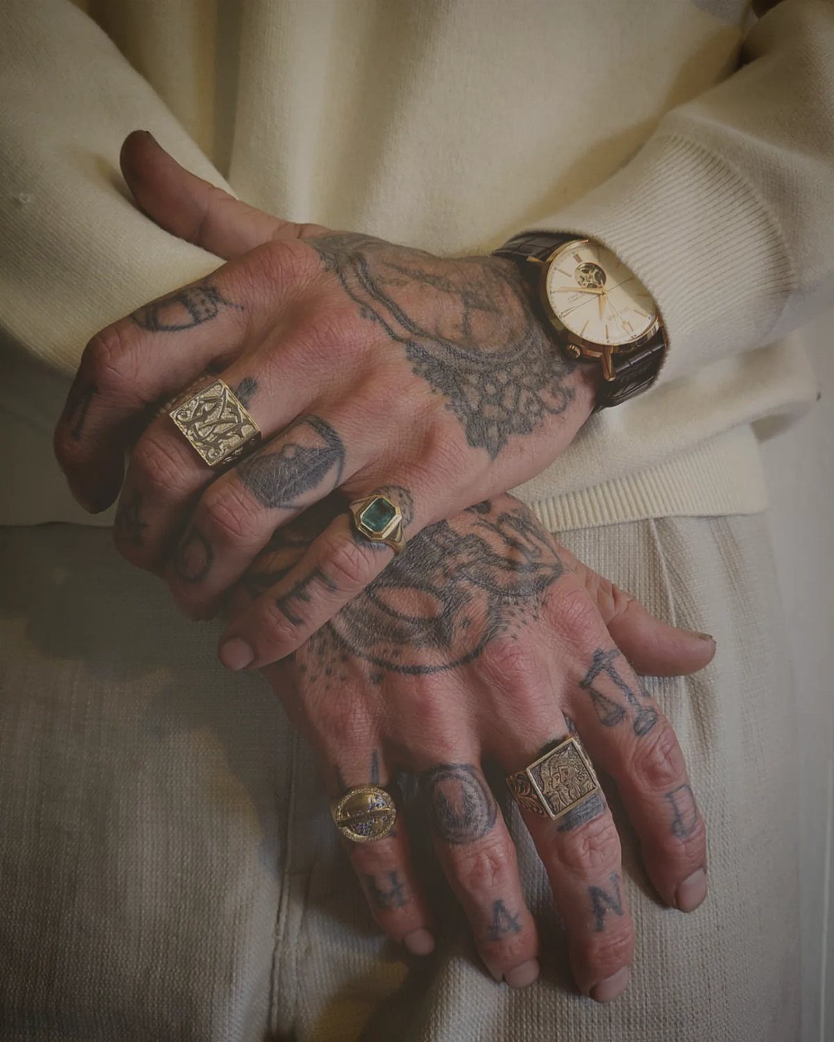 Tattooed hands wearing multiple gold signet and statement rings and a gold watch.