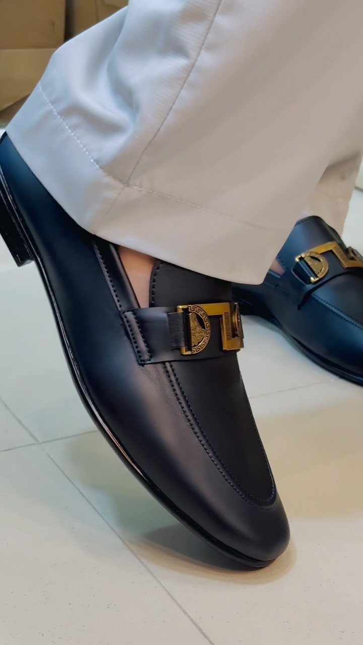Black leather loafers with gold buckles worn with light-colored trousers on white tiles.