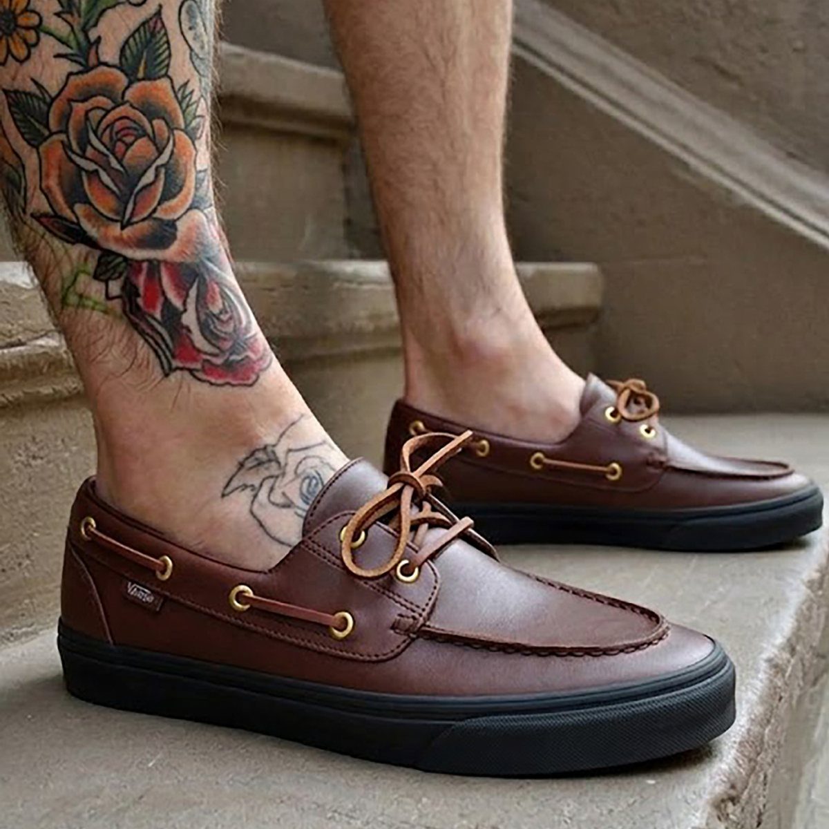 Brown leather deck shoes with black soles worn by a tattooed person.