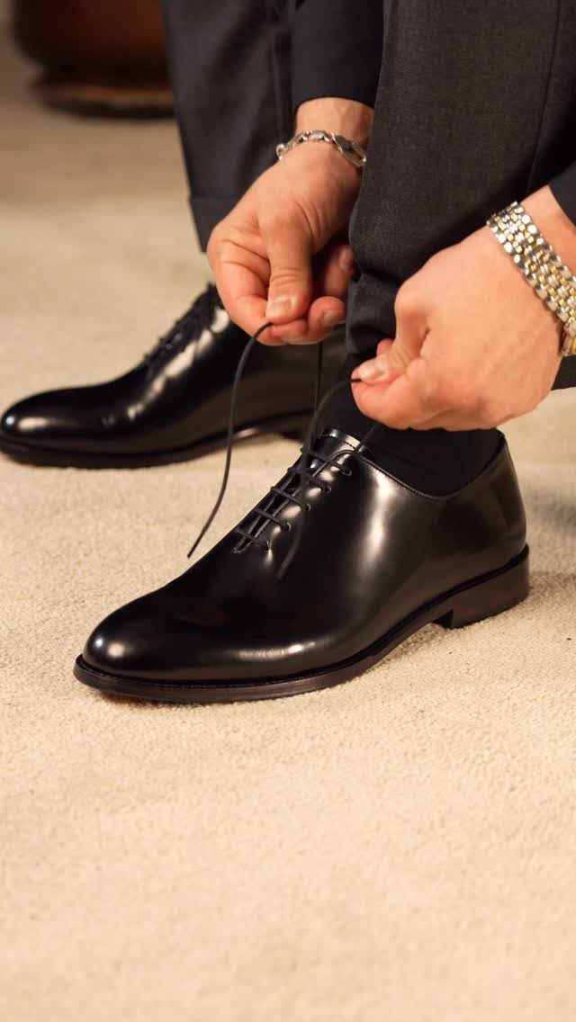Black leather Oxford shoes being tied by a man wearing dark trousers.