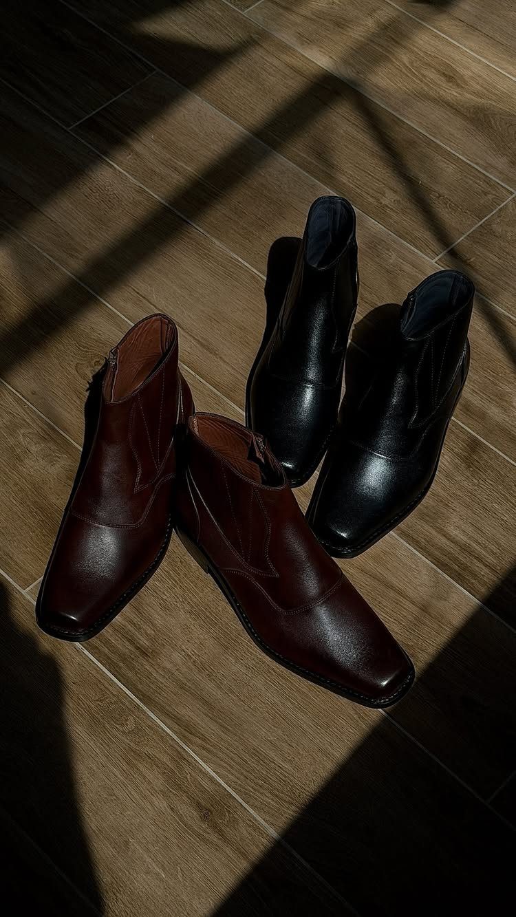 Brown and black leather dress boots resting on a light brown wooden floor.