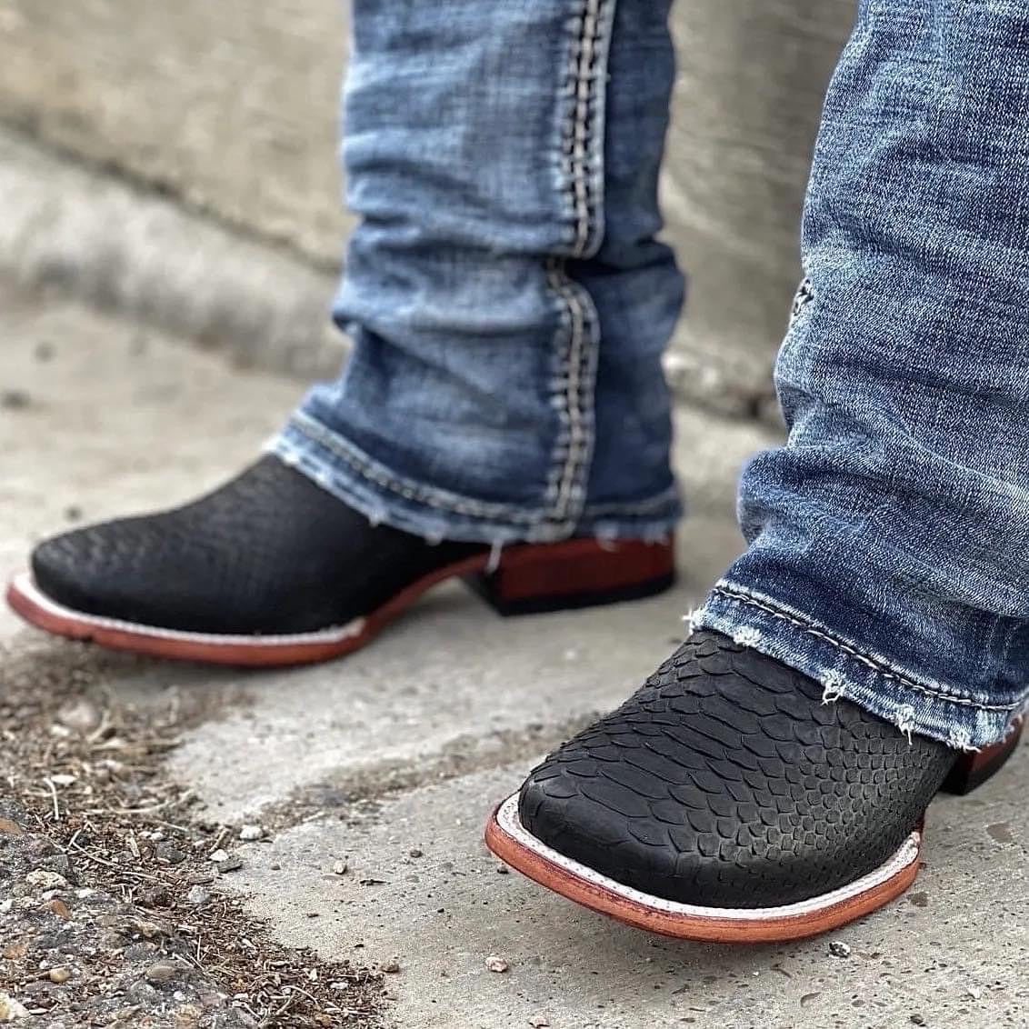 Black textured western boots with brown soles and blue denim jeans on concrete.