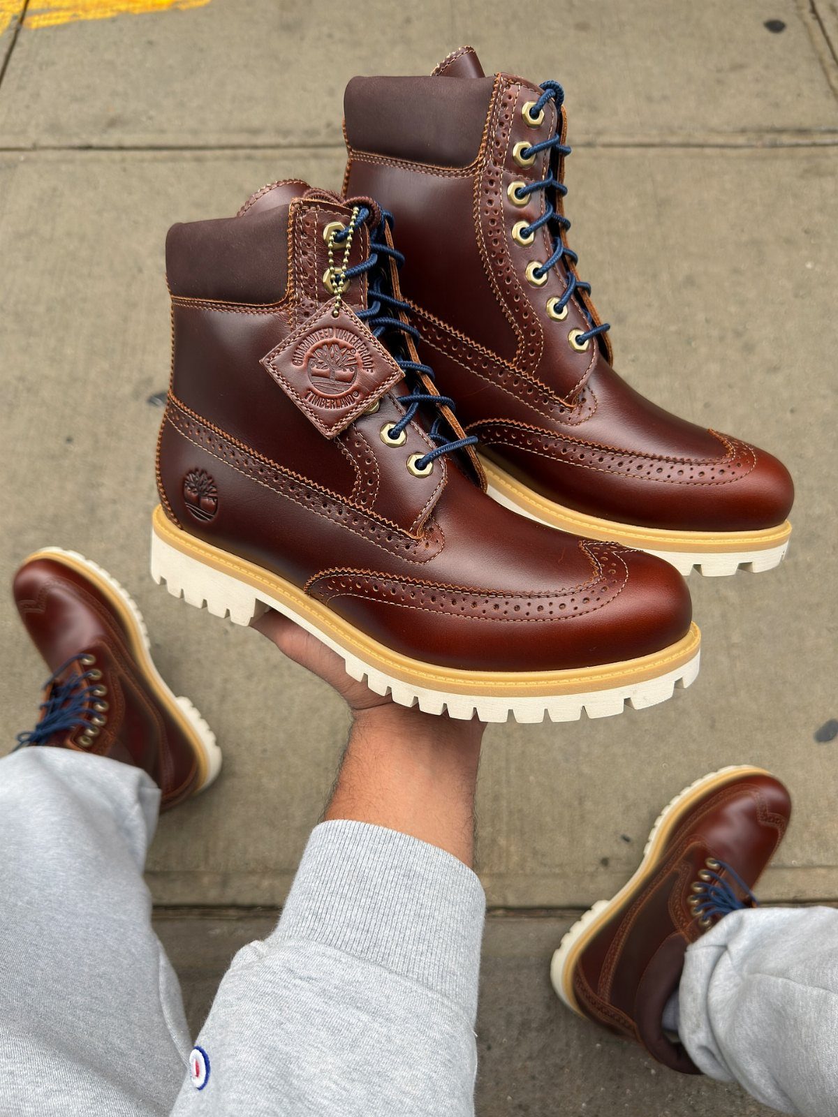 Brown leather brogue boots with blue laces and white soles held over grey pants.
