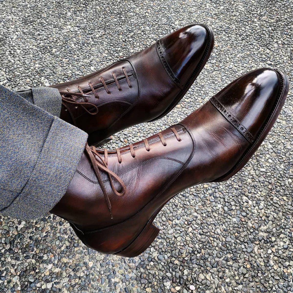 Brown leather captoe boots with grey trousers on a gravel ground.