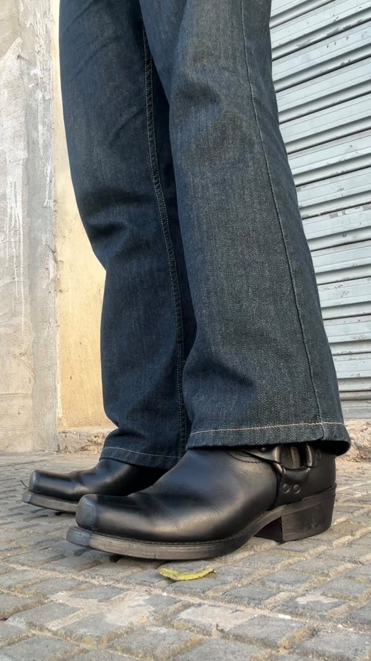 Black leather harness boots with straps worn with dark blue denim jeans.