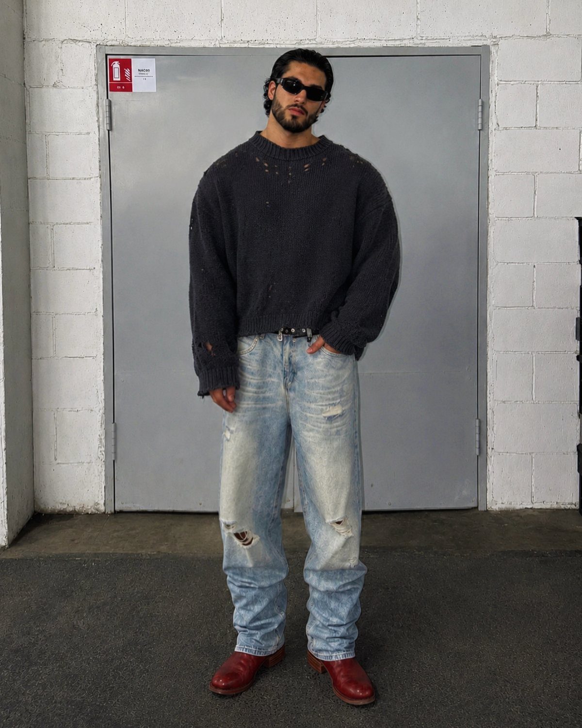 Man in oversized grey sweater and distressed jeans wearing reddish-brown leather boots.