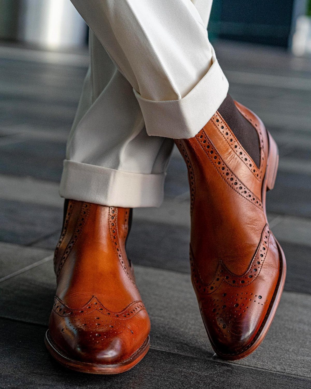 Tan leather wingtip boots with decorative brogueing and white cuffed trousers.
