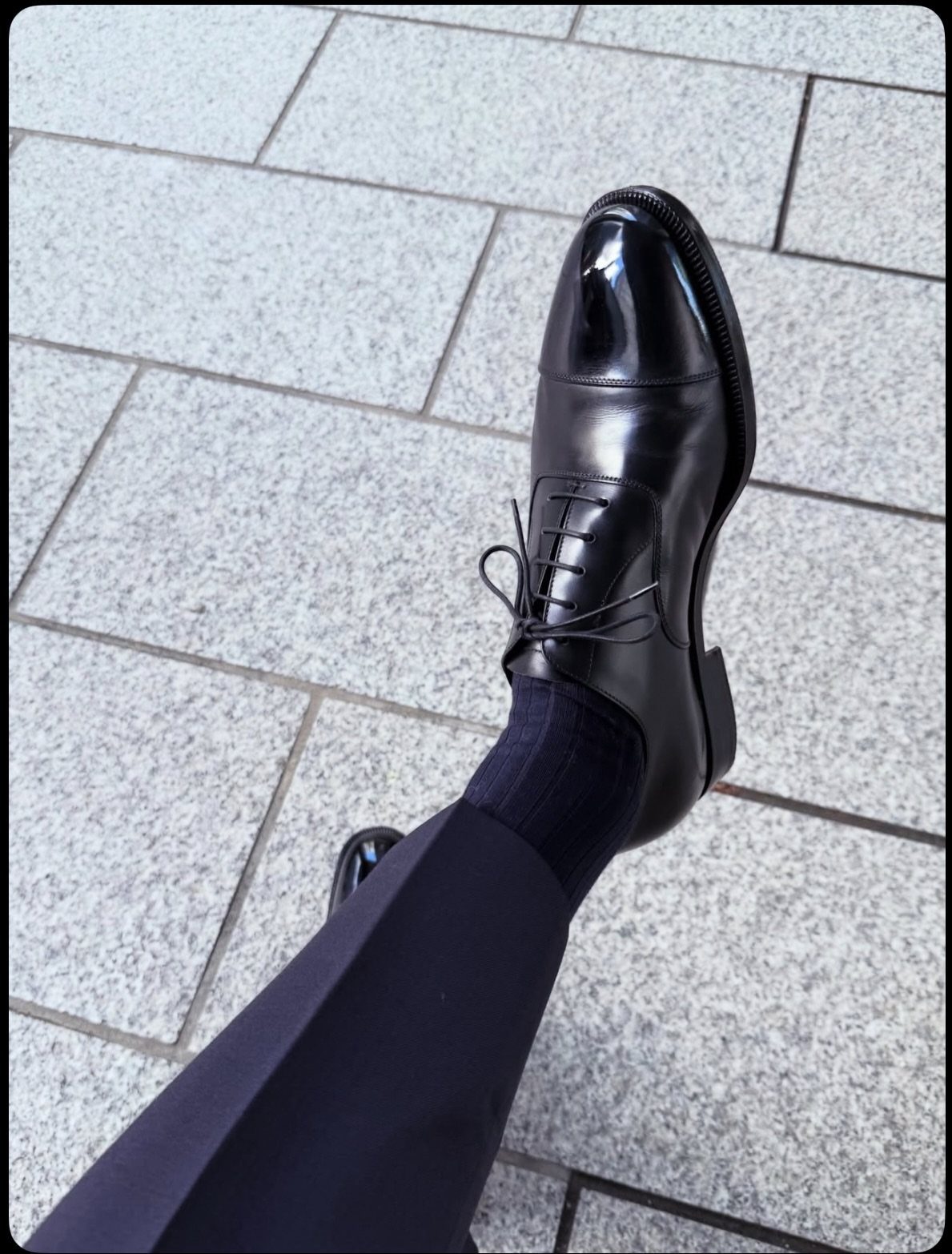 Polished black leather oxford shoes worn with navy trousers on stone tiles.