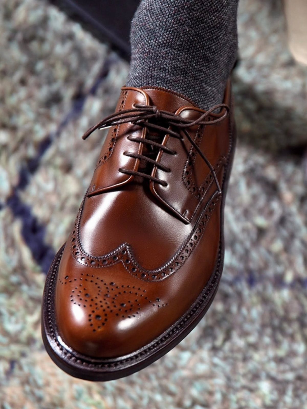 Brown leather wingtip derby shoe with grey socks on a stone background