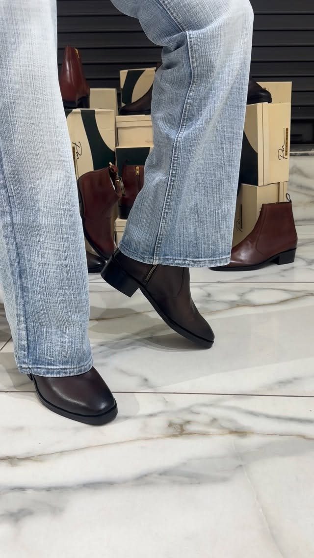 Dark brown leather Chelsea boots worn with light blue jeans on marble flooring.