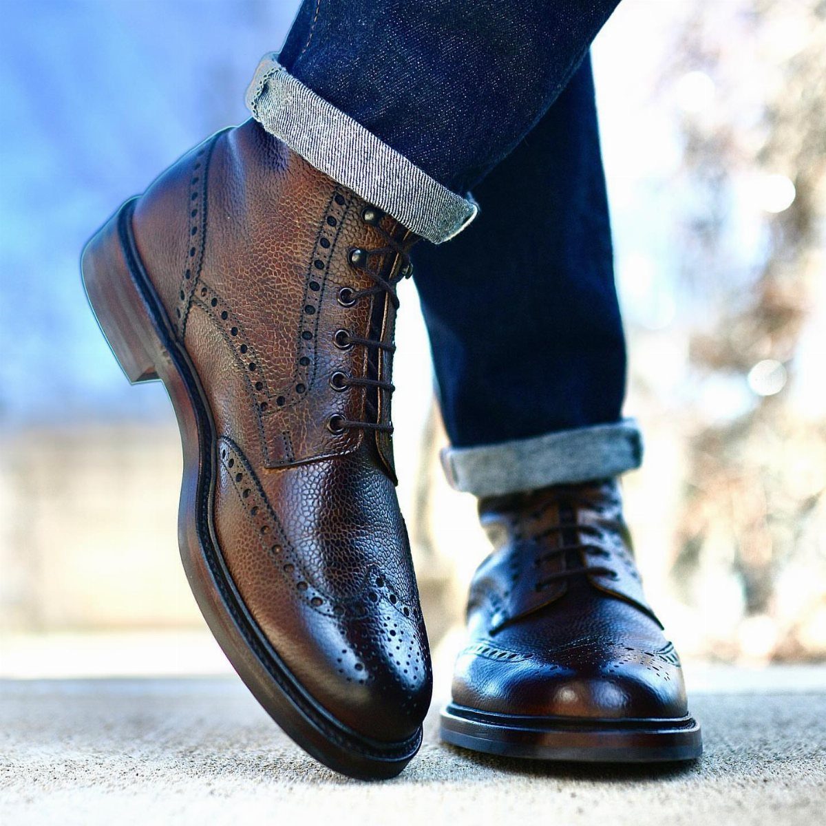 Brown leather wingtip boots with decorative perforations and cuffed dark blue jeans.