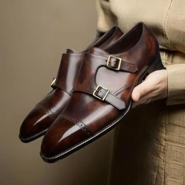 Brown leather double monk strap shoes with gold buckles held by a hand.